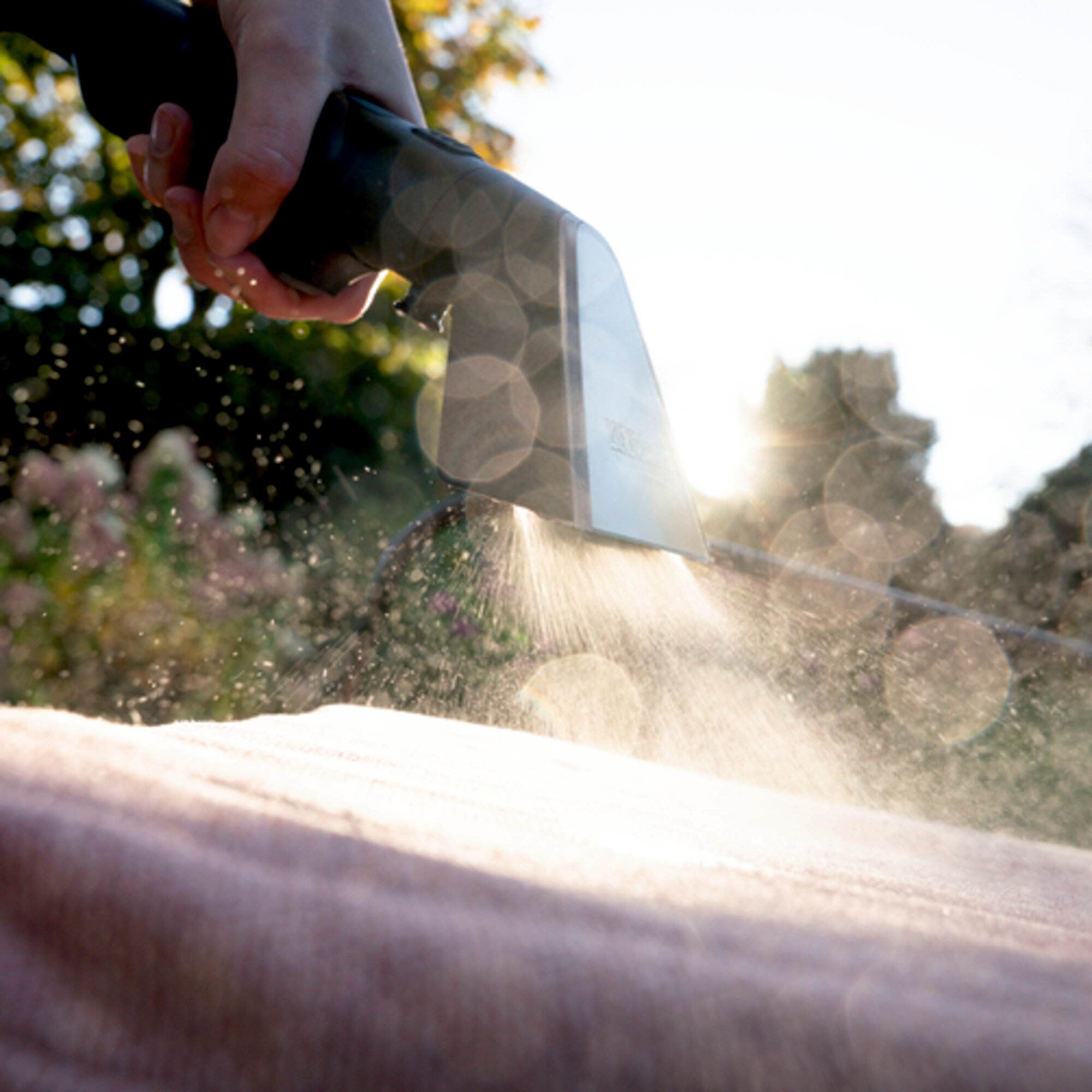 Dłoń trzymająca dyszę czyszczącą rozpylającą wodę na różową powierzchnię. Jasne słońce w tle.