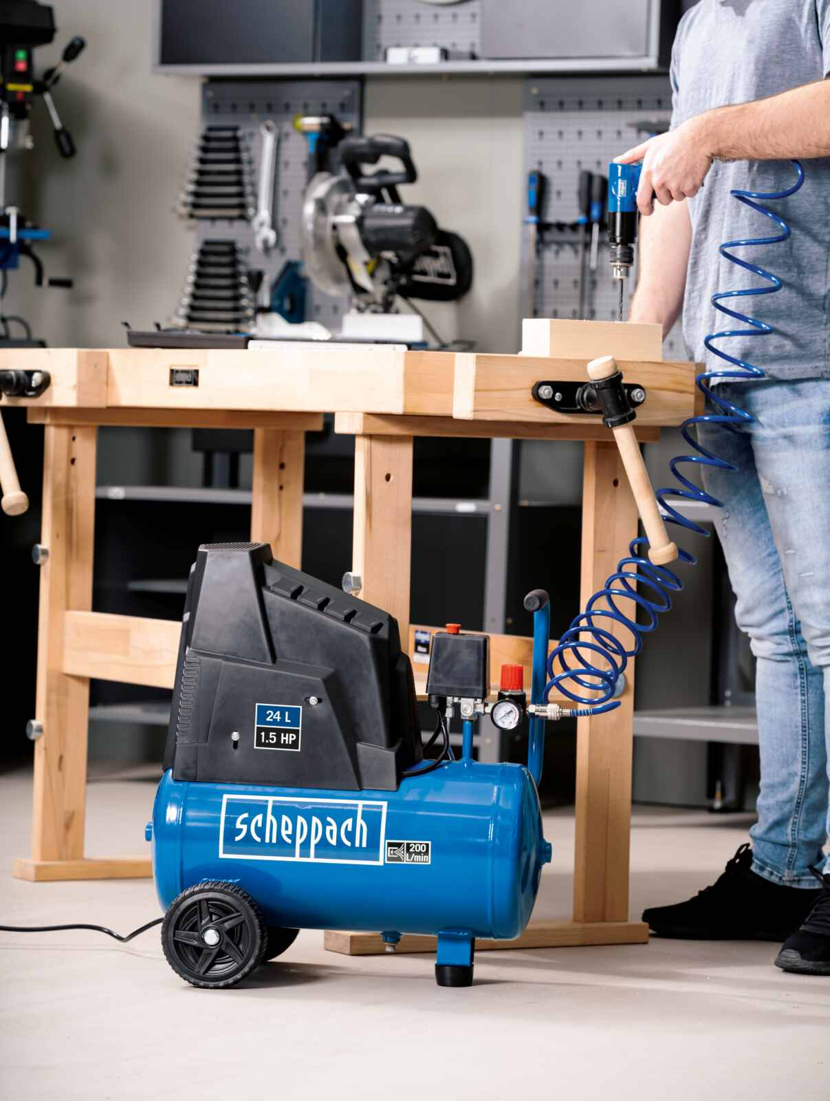 Man boort in hout aan een werkbank met gereedschap en een blauwe compressor.