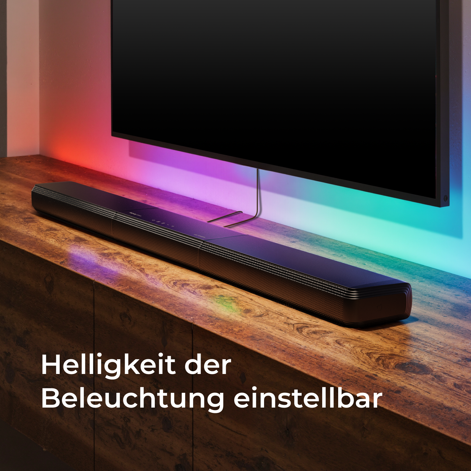 Eine schwarze Soundbar auf einem Holzschrank. Ein Fernseher ist darüber. Bunte Lichter hinter dem Fernseher. Text: Helligkeit einstellbar.