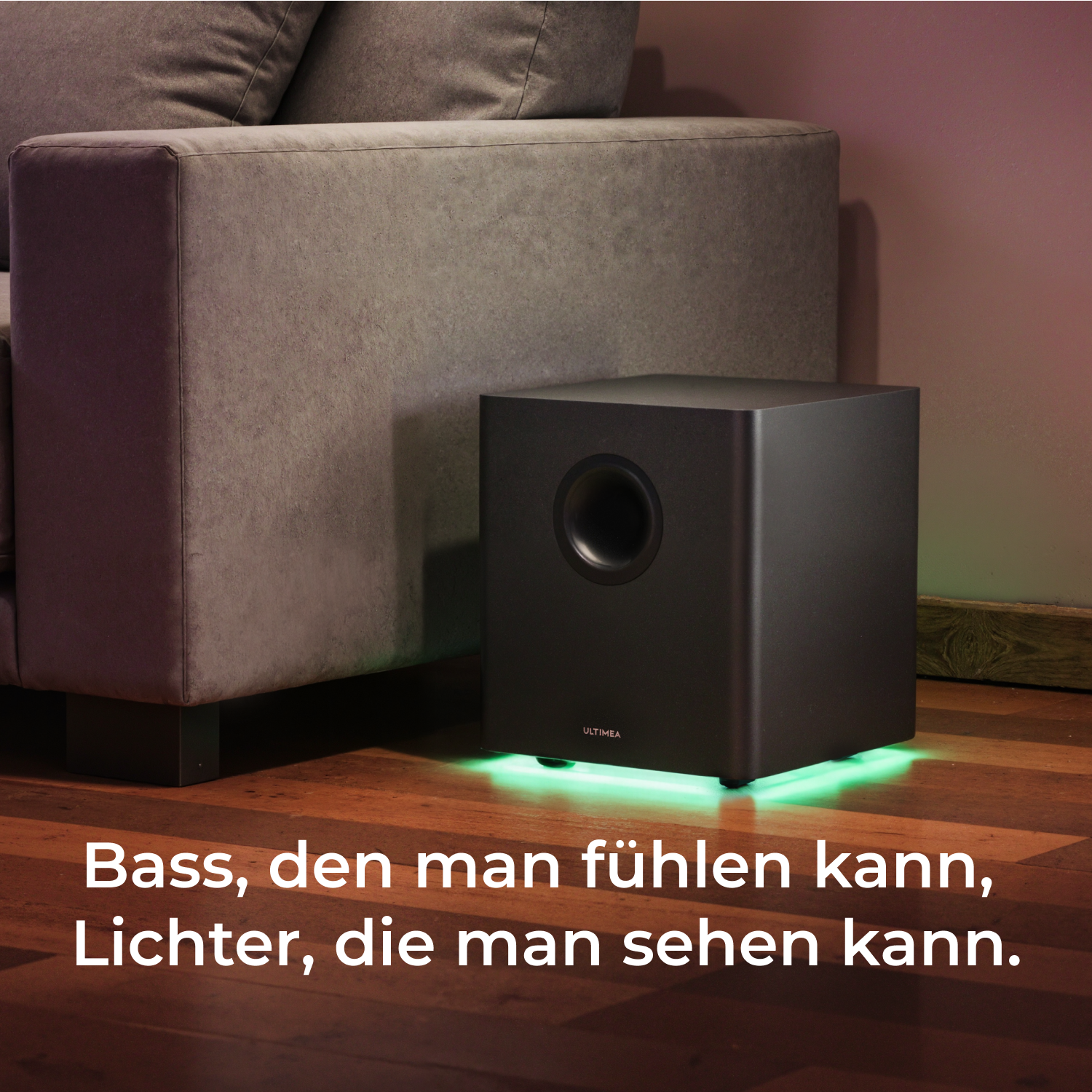 Schwarzer Subwoofer, der grünes Licht aussendet, in der Nähe eines grauen Sofas in einem Raum mit Holzboden.