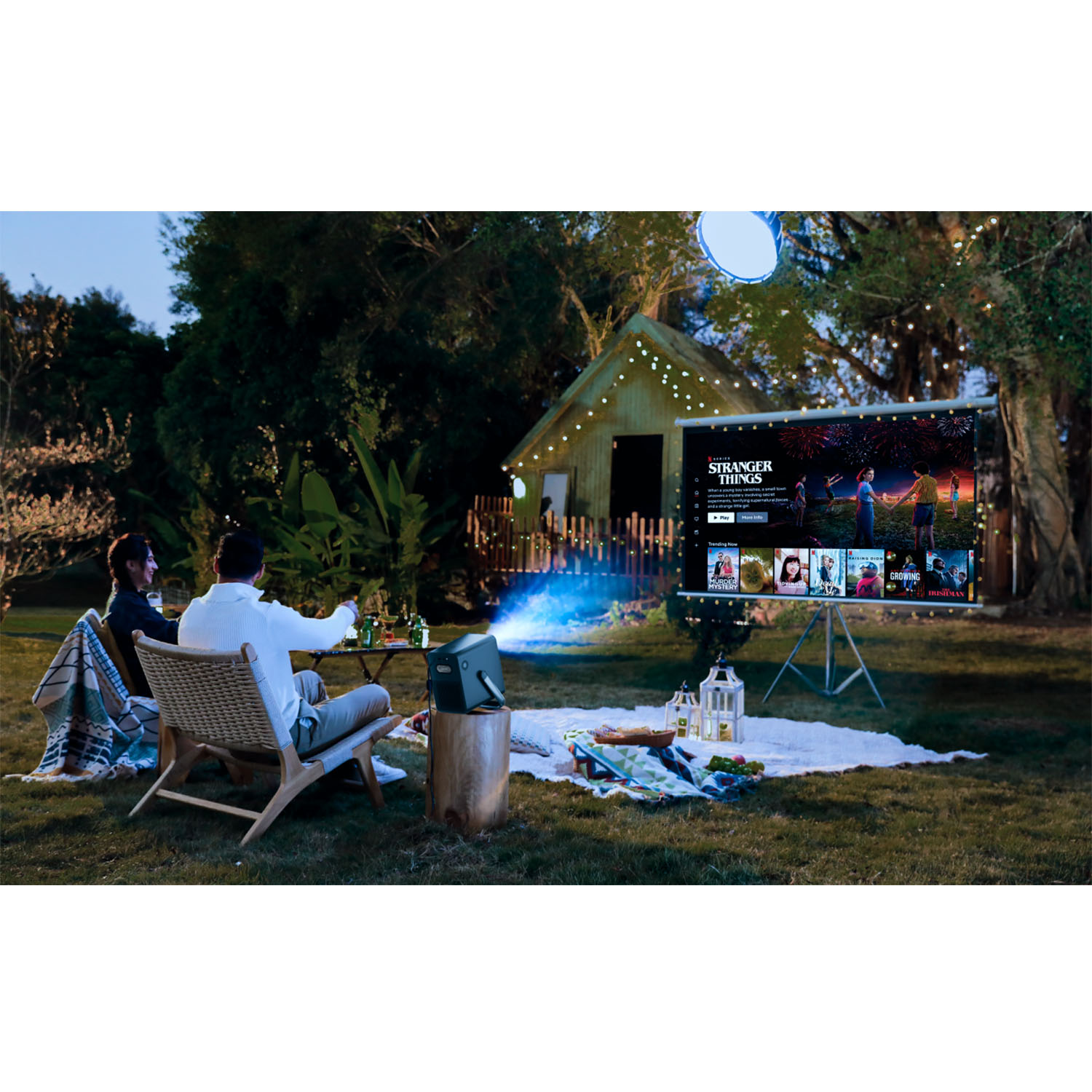 Leute schauen im Garten einen Film auf einer großen Leinwand. Ein Projektor ist vor ihnen, ein kleines Gebäude dahinter.