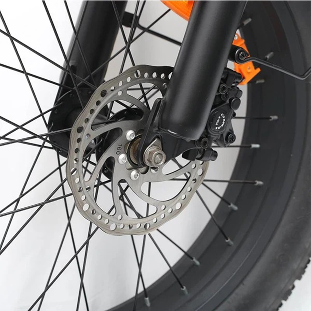 Primer plano de un freno de disco de bicicleta. El disco es plateado con agujeros, la pinza es negra. Neumático negro al fondo.