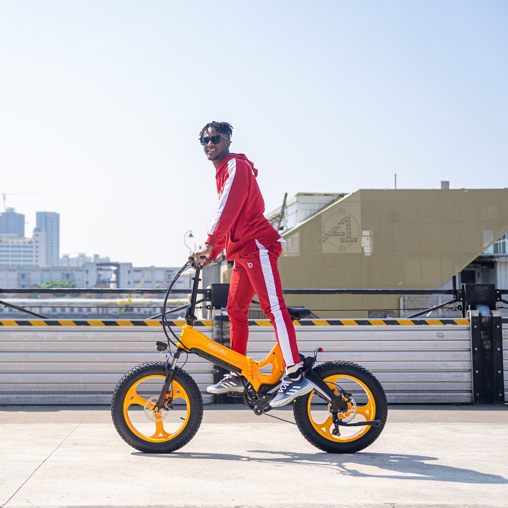 Ein Mann in roter Kleidung fährt auf einem orange-schwarzen E-Bike auf Beton, sonniger Tag.