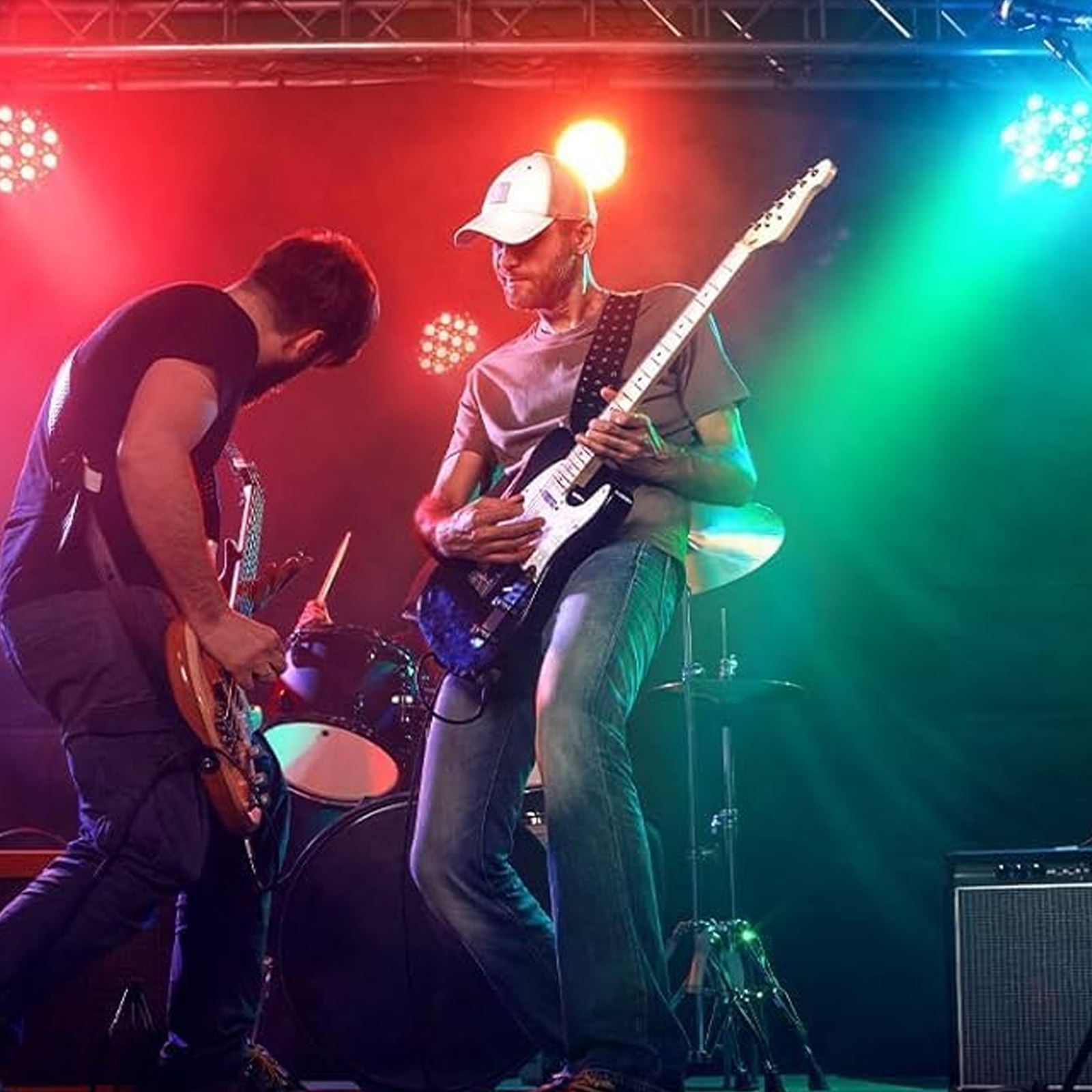 Zwei Männer spielen Gitarre auf einer Bühne mit Schlagzeug unter rotem und grünem Bühnenlicht.