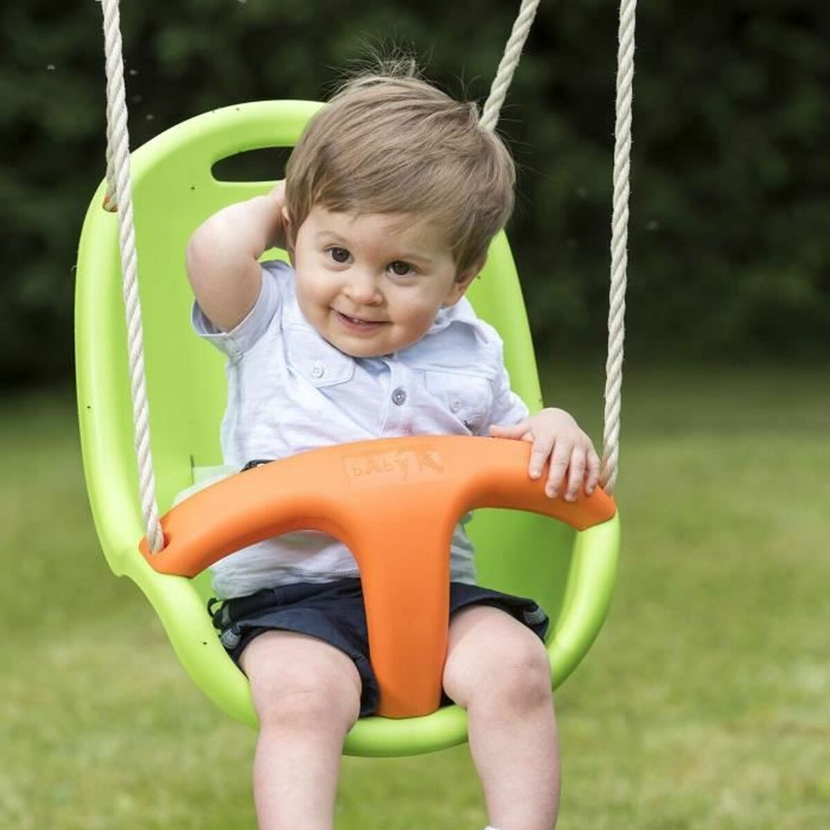 Un bambino sorridente seduto su un'altalena verde e arancione, con le mani sulla testa.