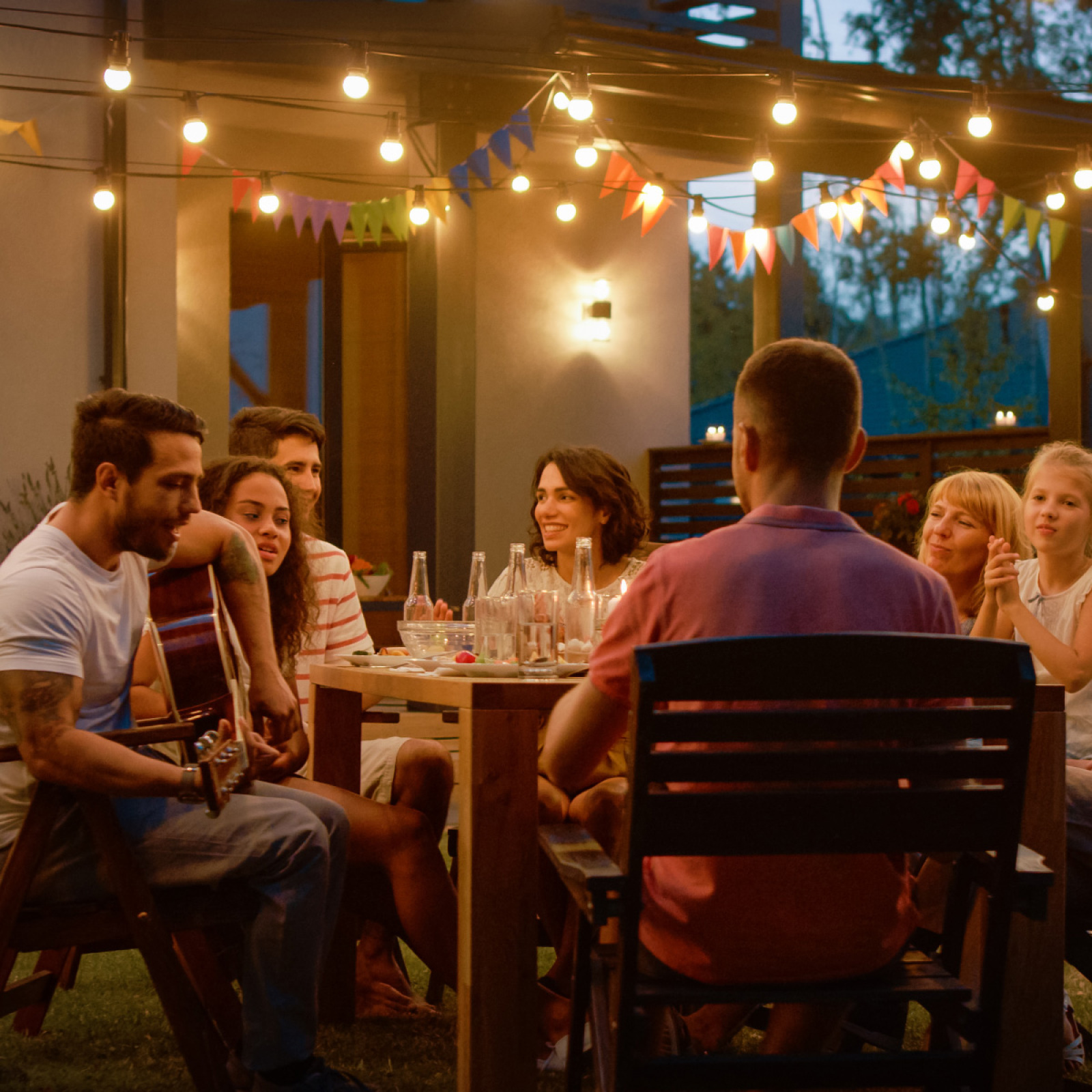 Personengruppe bei einer Gartenparty, einige spielen Gitarre. Lichterketten beleuchten die Szene.