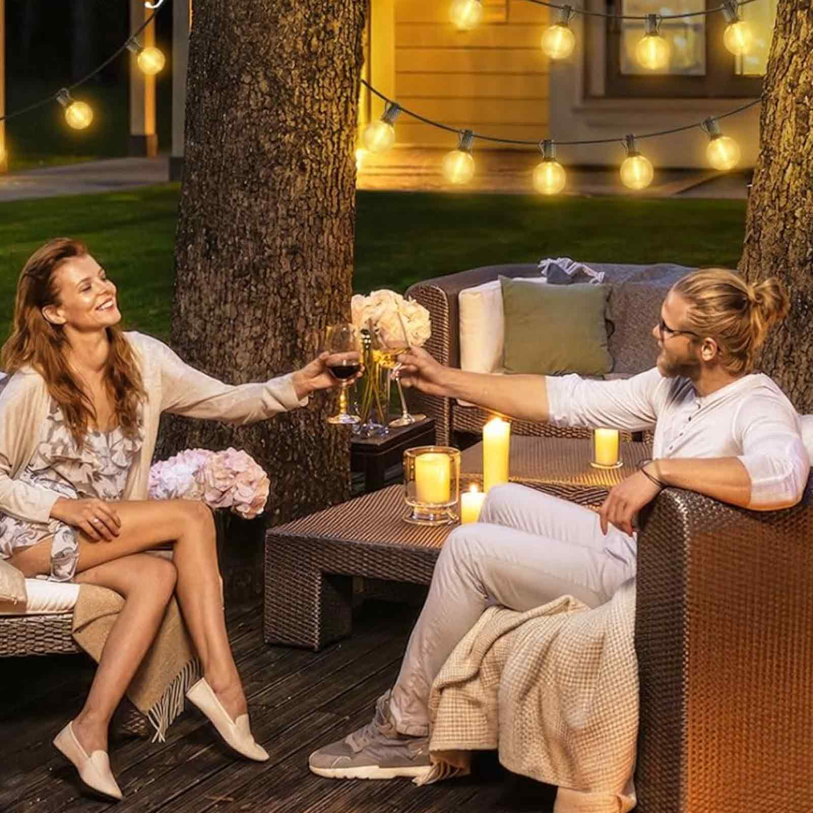 Ein Paar stößt mit Wein an, sitzt draußen mit Lichterketten und Kerzen auf einem Tisch.