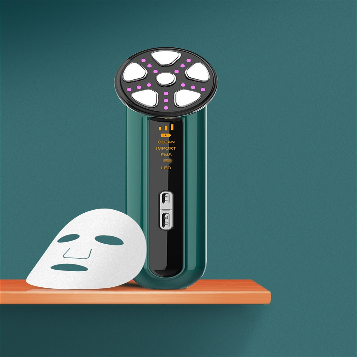Grünes Gesichtsmassagegerät mit LED-Leuchten und einer Maske auf einem Regal vor einer türkisfarbenen Wand.