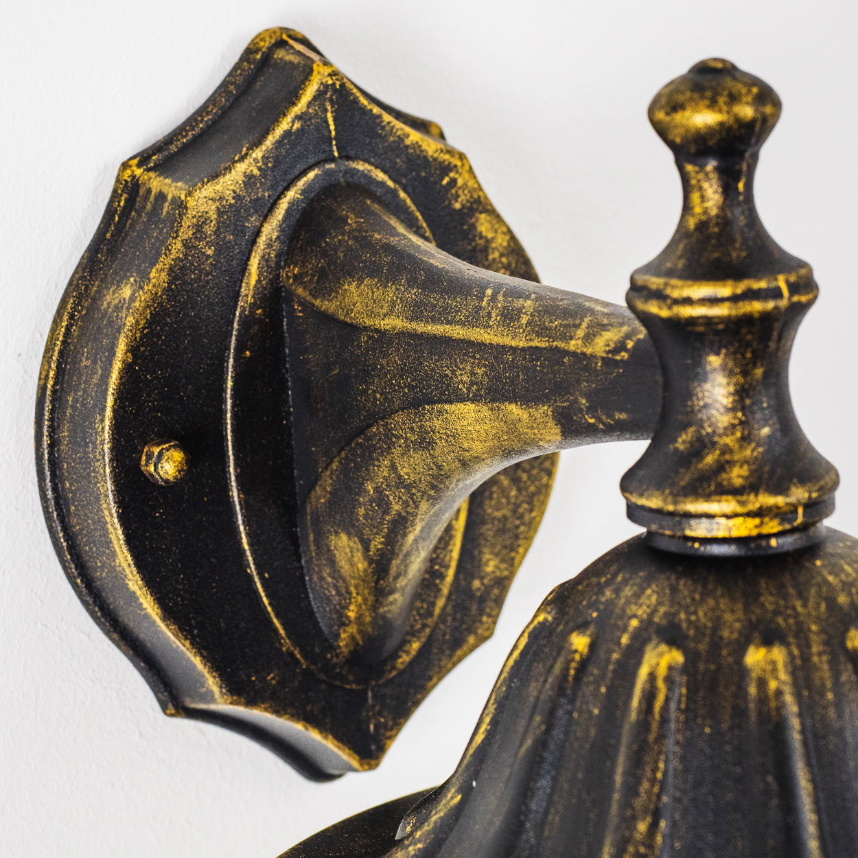 Nahaufnahme einer verzierten Wandlampe, schwarz und gold. Zeigt die Halterung. Weißer Hintergrund.