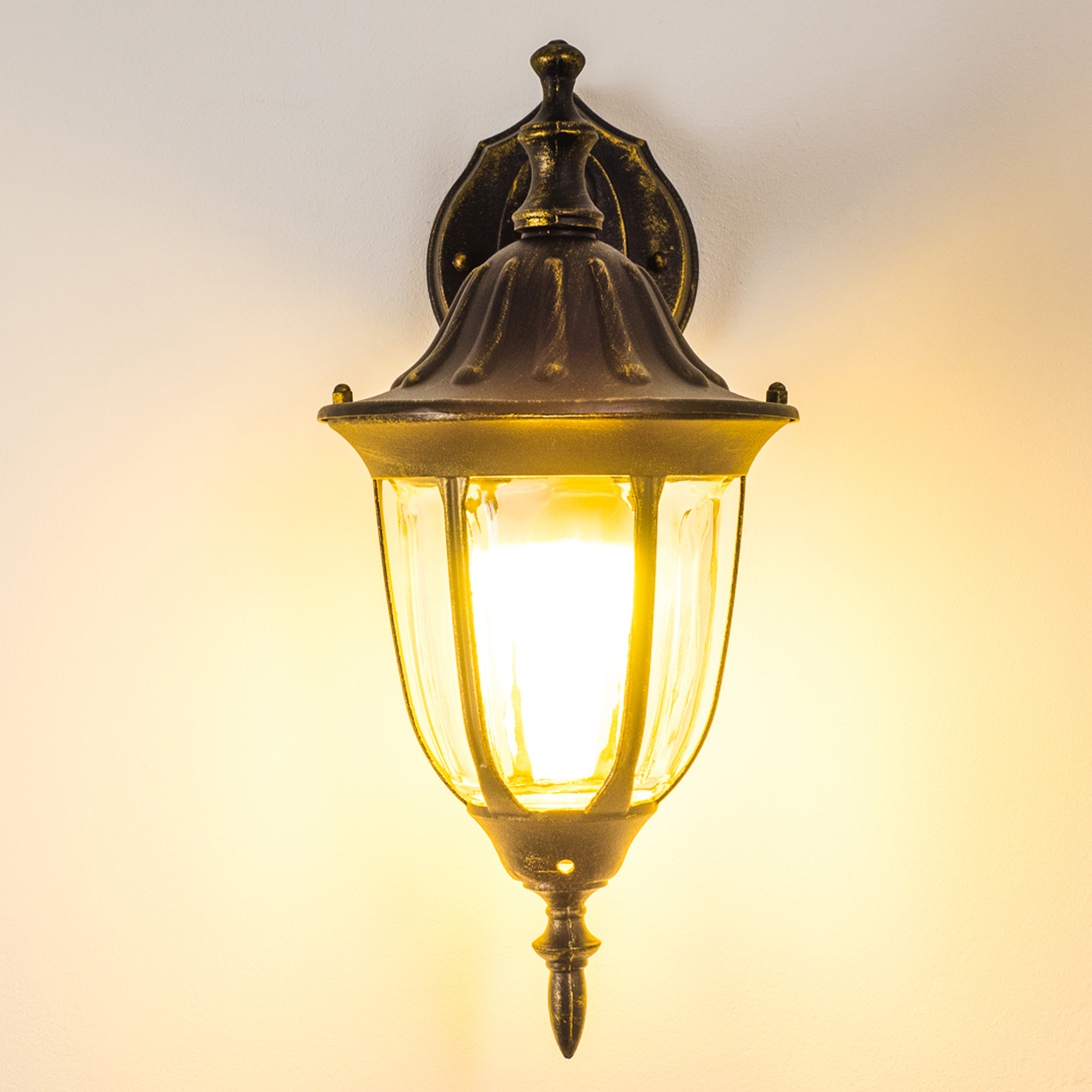 Bronze-farbene Wandlampe mit hellem Licht, an einer weißen Wand montiert. Die Lampe ist klassisch im Design.