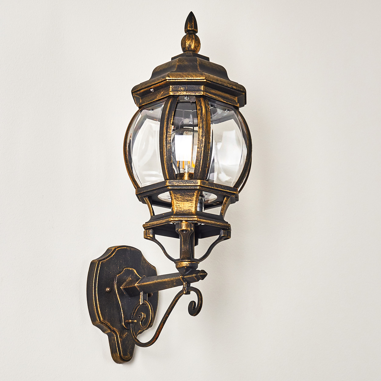 Aufwendige bronzefarbene Wandlampe mit klaren Glasscheiben, an einer weißen Wand. Detailliertes Design, helles Licht.