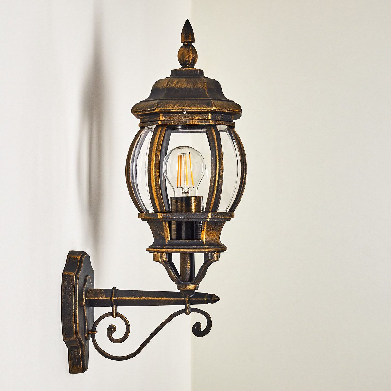 Aufwendige bronzefarbene Wandlampe mit klaren Glasscheiben, an einer weißen Wand. Detailliertes Design, helles Licht.