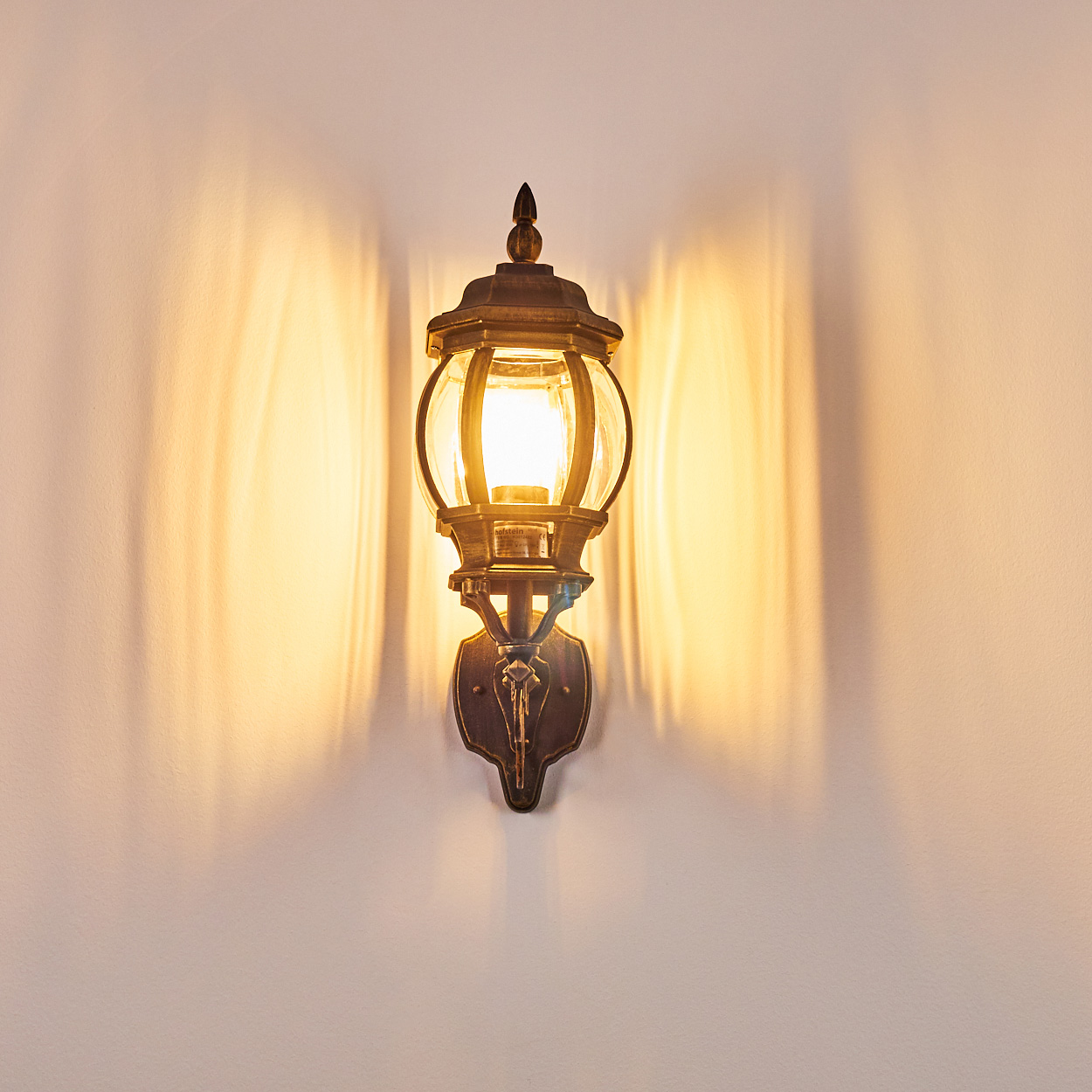 Bronze-Wandlampe, die warmes Licht aussendet und Schatten wirft. Klassisches Design, an weißer Wand.
