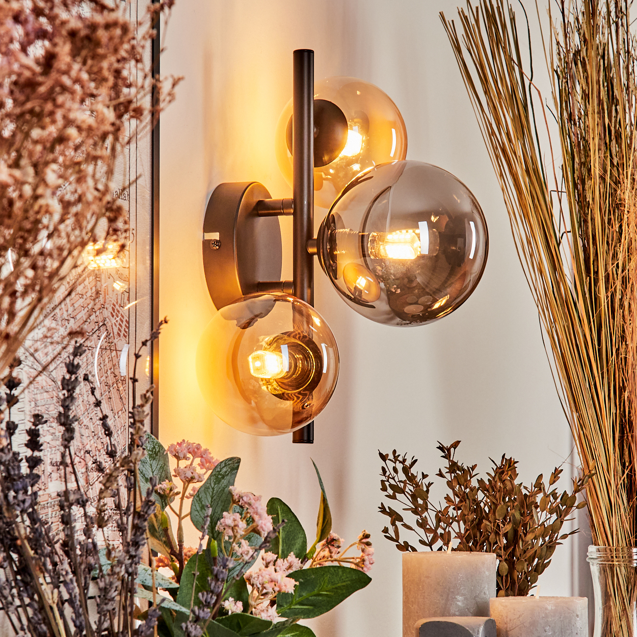 Wandlampe mit drei Glaskugeln, bernsteinfarben und grau, auf schwarzem Rahmen. Trockenpflanzen im Hintergrund.