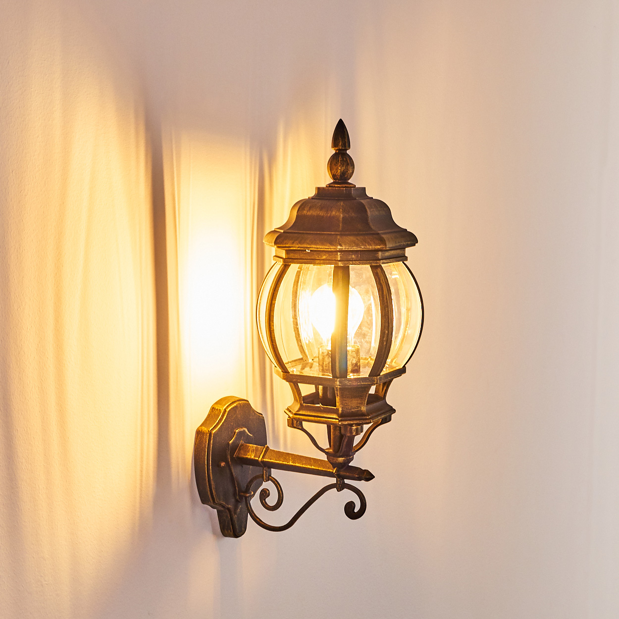 Bronze Wandlampe mit klarem Glasschirm, eingeschaltet, wirft Licht auf die Wand.