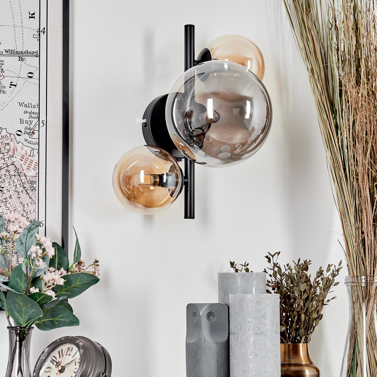 Wandlampe mit drei Glaskugeln. Zwei bernsteinfarben, eine klar. Schwarzer Arm. Hintergrund: Karte, Pflanzen, Kerzen, Uhr.