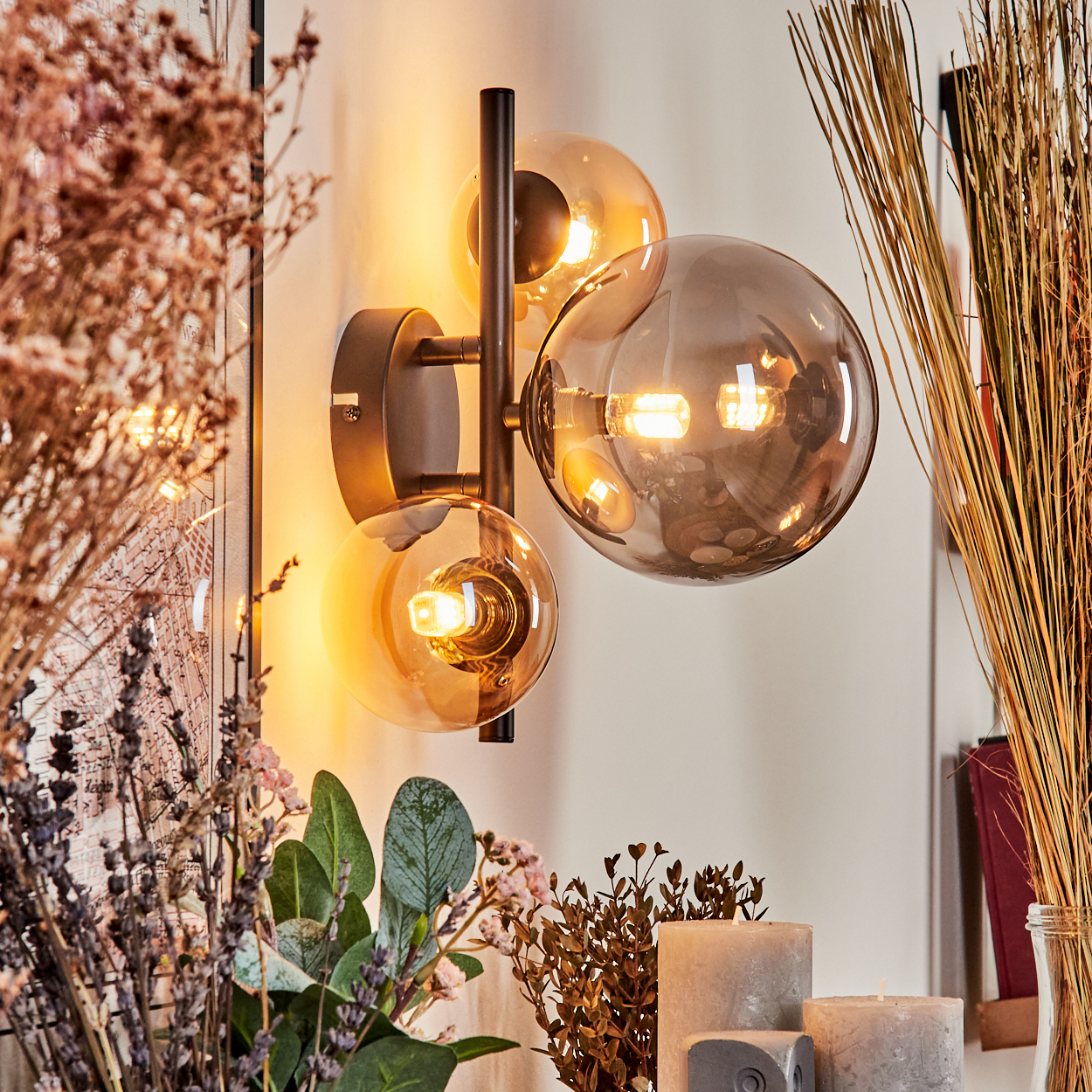 Wandlampe mit drei Glaskugeln. Zwei bernsteinfarben, eine klar. Schwarzer Arm. Hintergrund: Blumen, Kerzen, trockene Pflanzen.