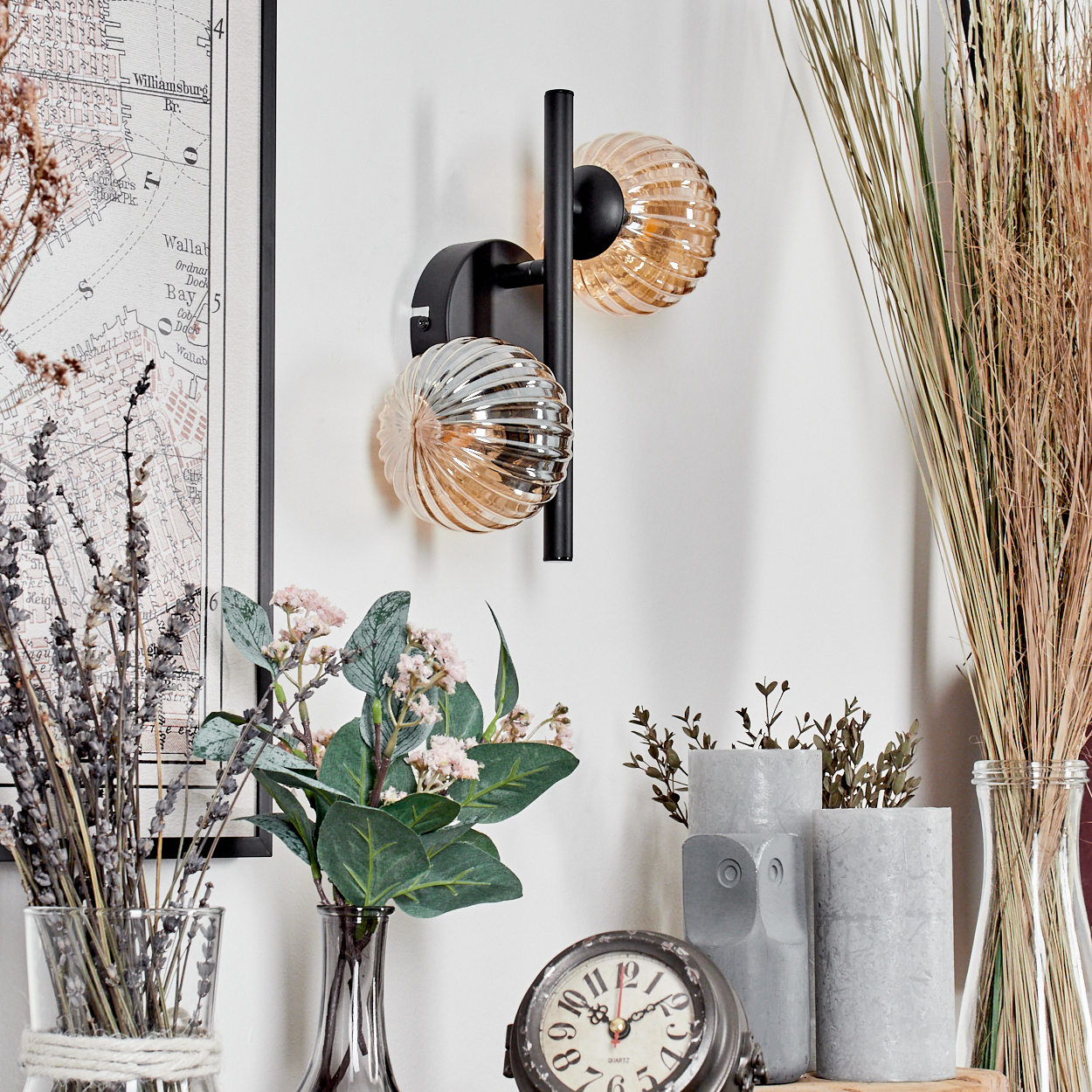 Wandleuchte mit drei Glaskugeln, schwarzem Rahmen. Blumen, Uhr und Karte im Hintergrund.