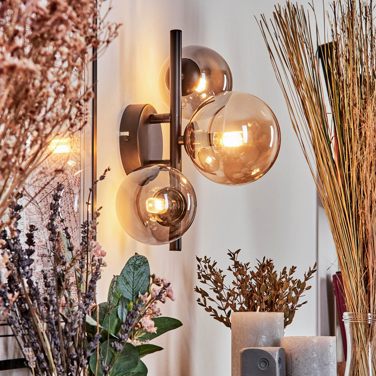 Eine Wand mit einer Lampe mit drei Glaskugeln, umgeben von Pflanzen und Kerzen.