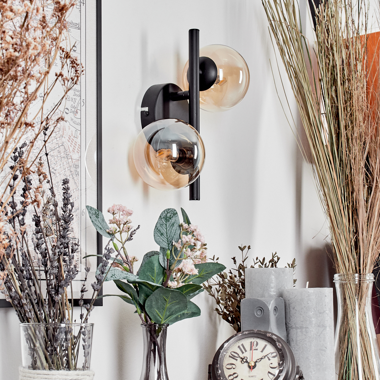 Wandleuchte mit zwei bernsteinfarbenen Glaskugeln, schwarzem Rahmen, mit Deko wie Blumen und Uhr.