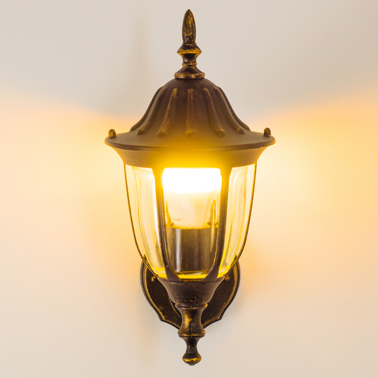 Bronze-Außenwandleuchte mit klarem Glas und einer brennenden Glühbirne. Sie ist an einer weißen Wand montiert.
