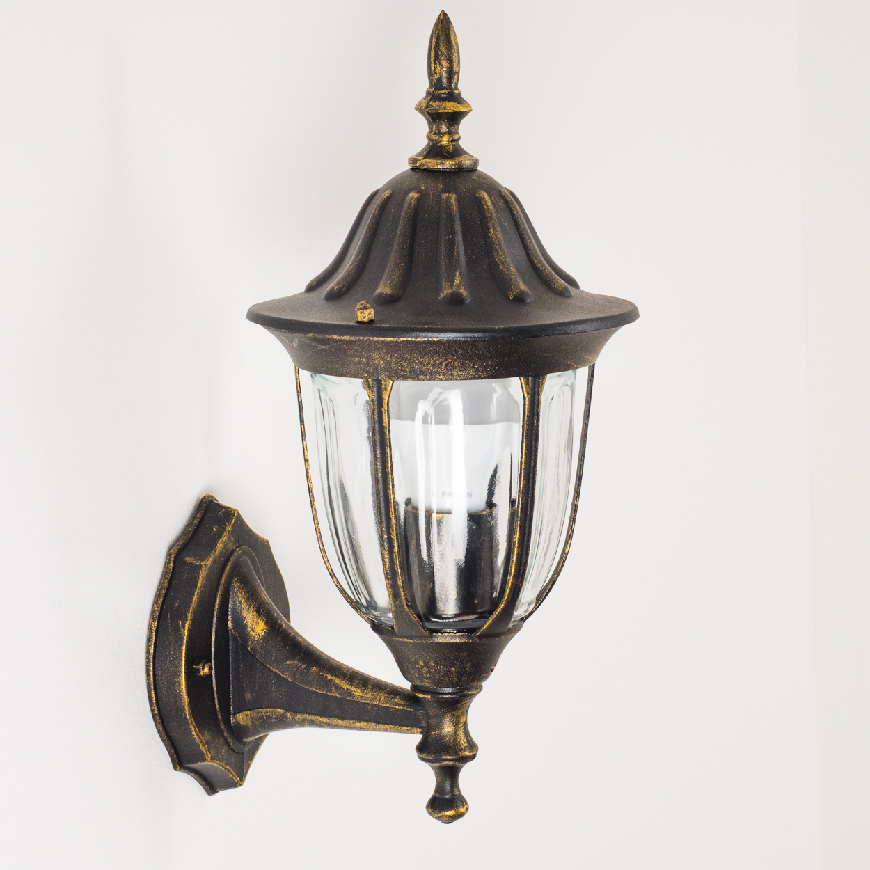 Verzierte Wandleuchte mit schwarz-goldener Oberfläche, an einer weißen Wand.