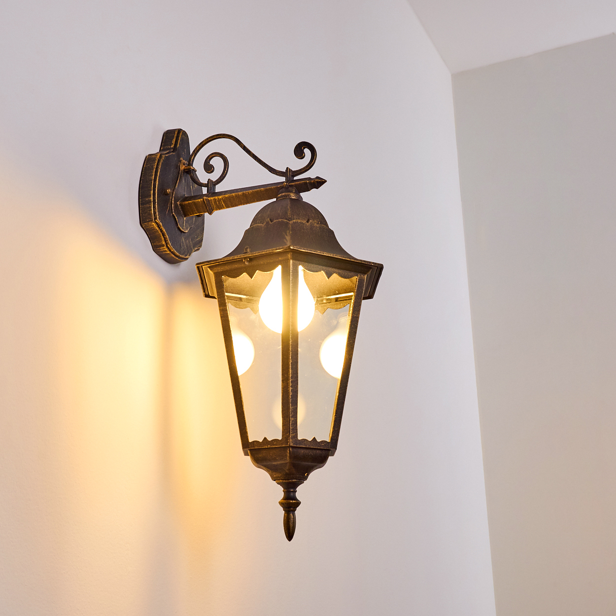 Verzierte schwarz-goldene Wandleuchte an einer weißen Wand, beleuchtet mit warmem Licht.