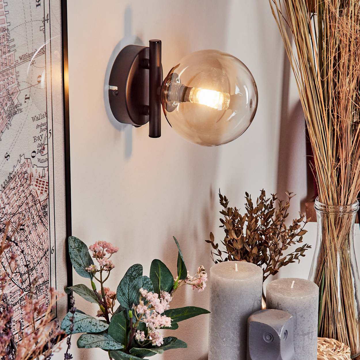Wandlampe mit brauner Glaskugel, Kerzen, Karte und Pflanzen. Der Hintergrund ist eine Wand.