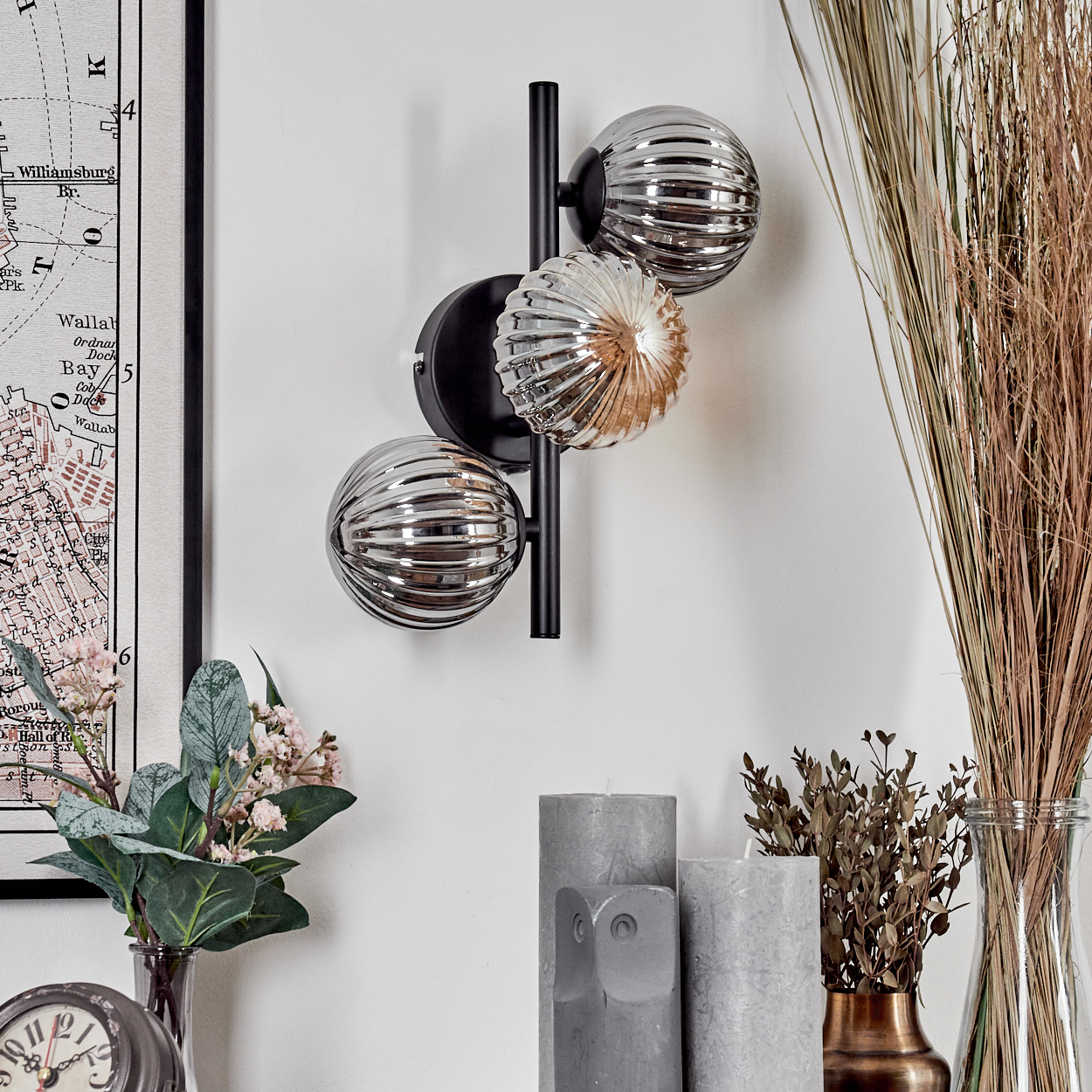 Wandlampe mit drei Glaskugeln, Kerzen und Blumen vor einer weißen Wand.