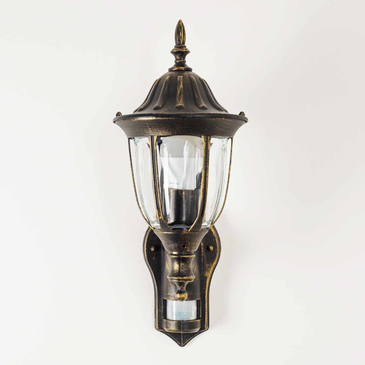 Verzierte Bronze-Wandlampe an weißer Wand. Klarglasschirm.