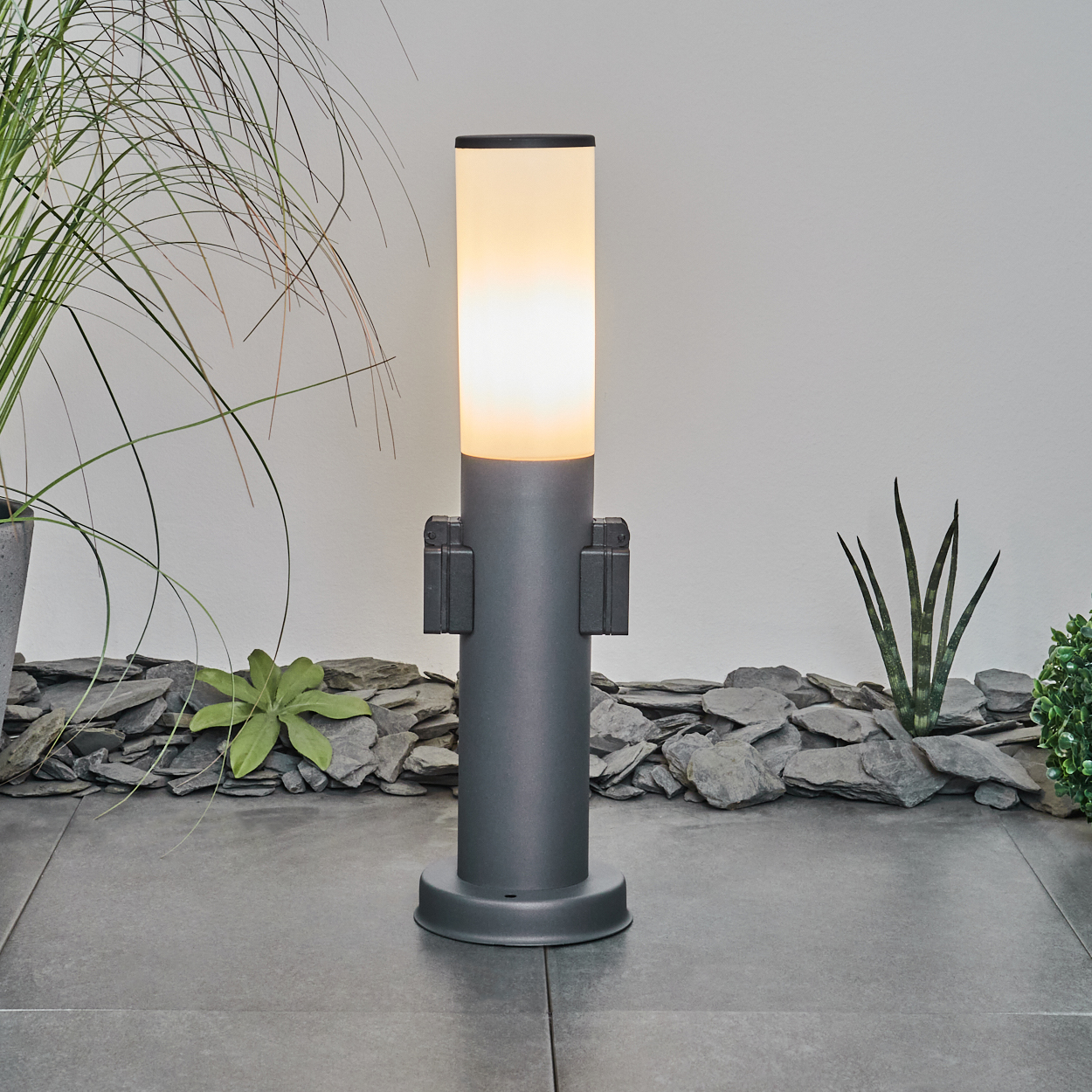 Beleuchtete graue Außenlampe neben Pflanzen und Steinen auf einer grauen Terrasse. Weiße Wand.