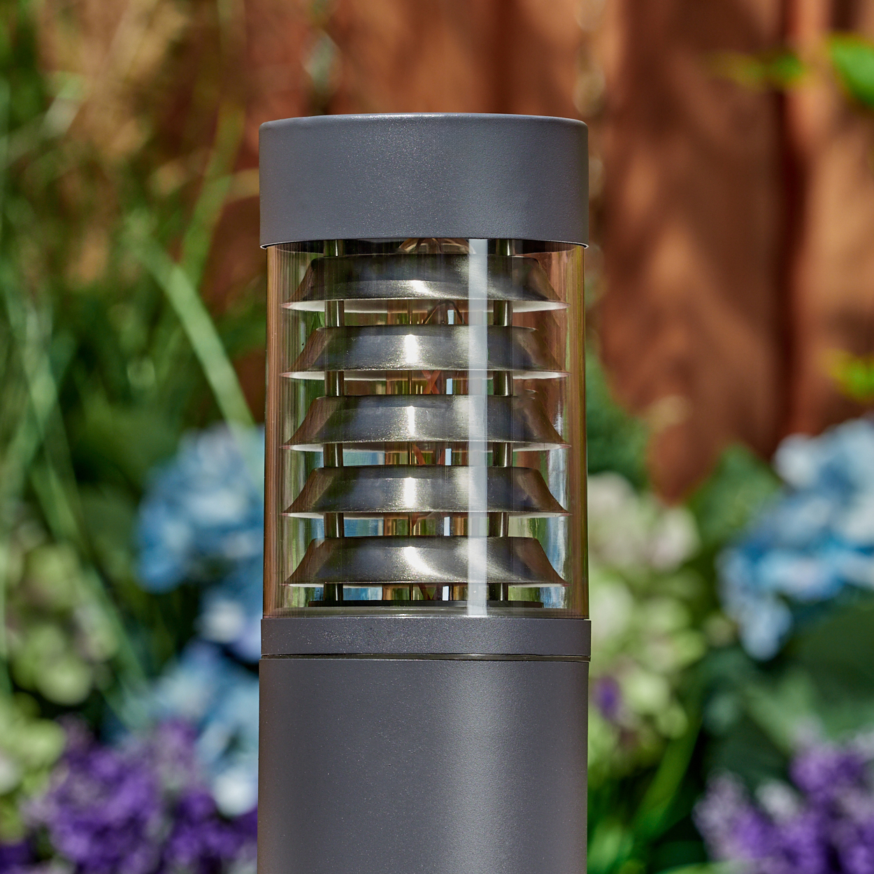 Eine graue zylindrische Außenlampe mit horizontalen Metalllamellen, mit verschwommenem floralen Hintergrund.