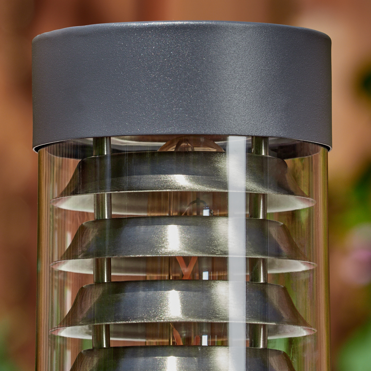 Nahaufnahme der grauen zylindrischen Außenlampe mit horizontalen Metalllamellen. Der Hintergrund ist unscharf.