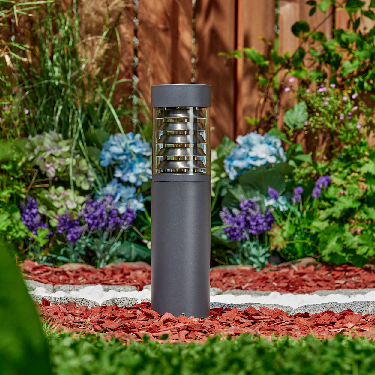 Dunkelgraue Außenlampe in einem Garten mit roter Mulch, Blumen und einem Holzzaun.