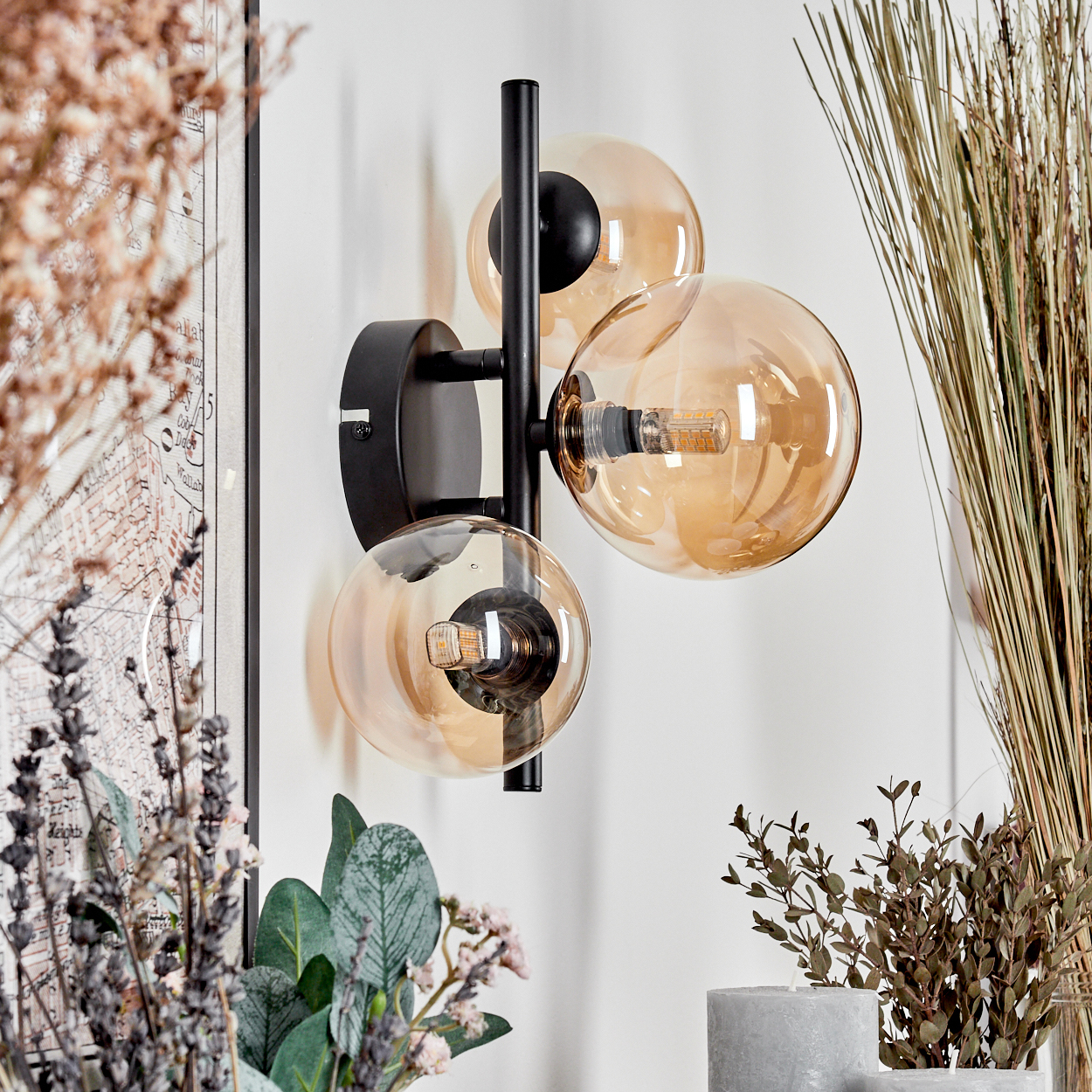 Schwarze Wandlampe mit drei bernsteinfarbenen Glaskugeln, weißer Wandhintergrund, getrocknete Pflanzen.