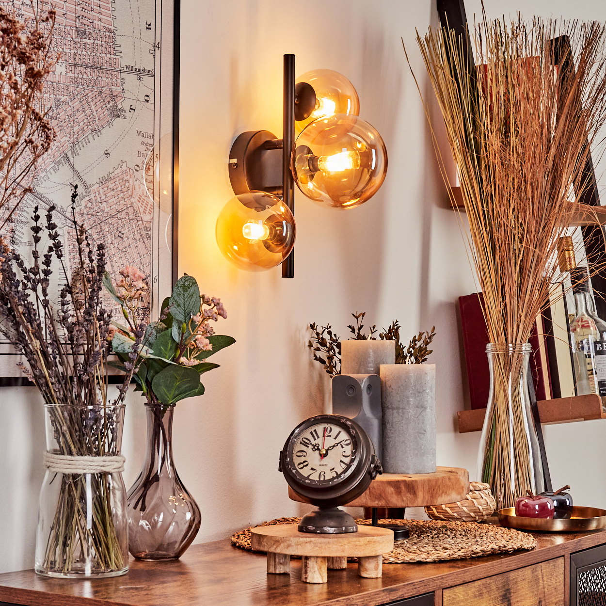Eine Wand mit einer schwarzen Lampe mit bernsteinfarbenen Glasschirmen, Kerzen, Vasen, Uhr, Karte und Pflanzen auf einem Holztisch.