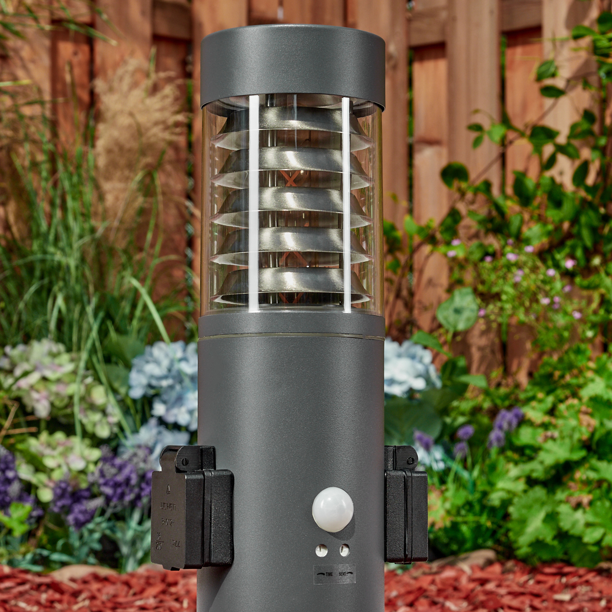 Eine graue Außenlampe mit offenem Deckel. Sie hat einen Bewegungssensor und steht in einem Garten mit roter Mulch.