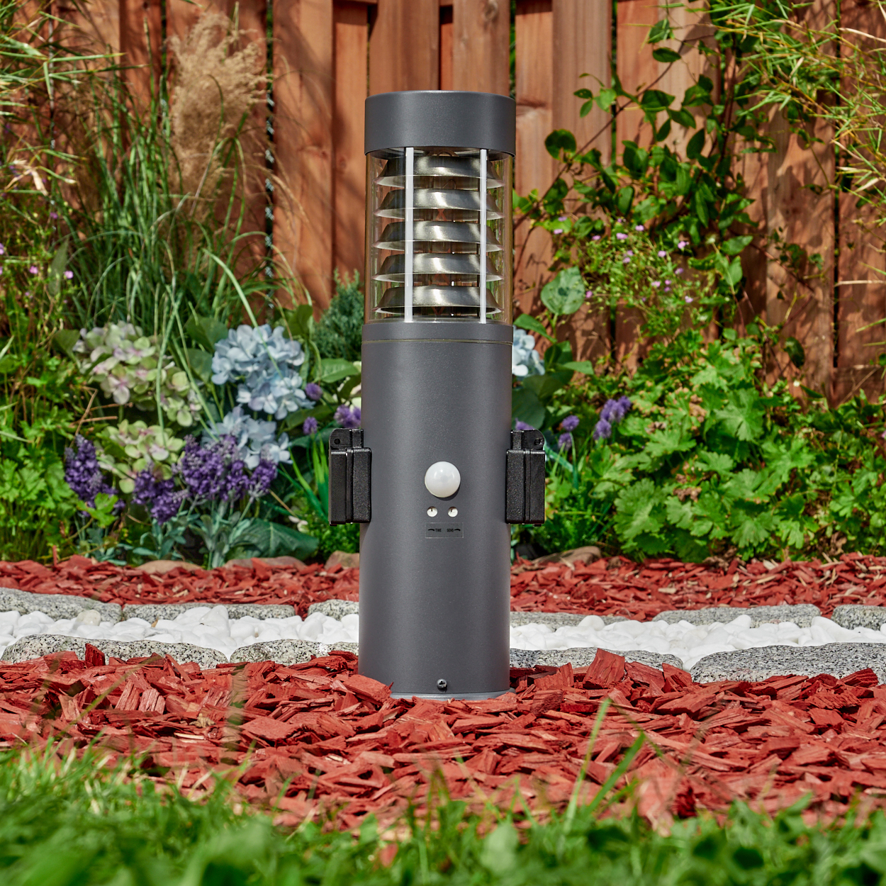 Eine graue Außenlampe mit Bewegungsmelder, aufgestellt in einem Garten mit roter Mulch und Holzzaun.