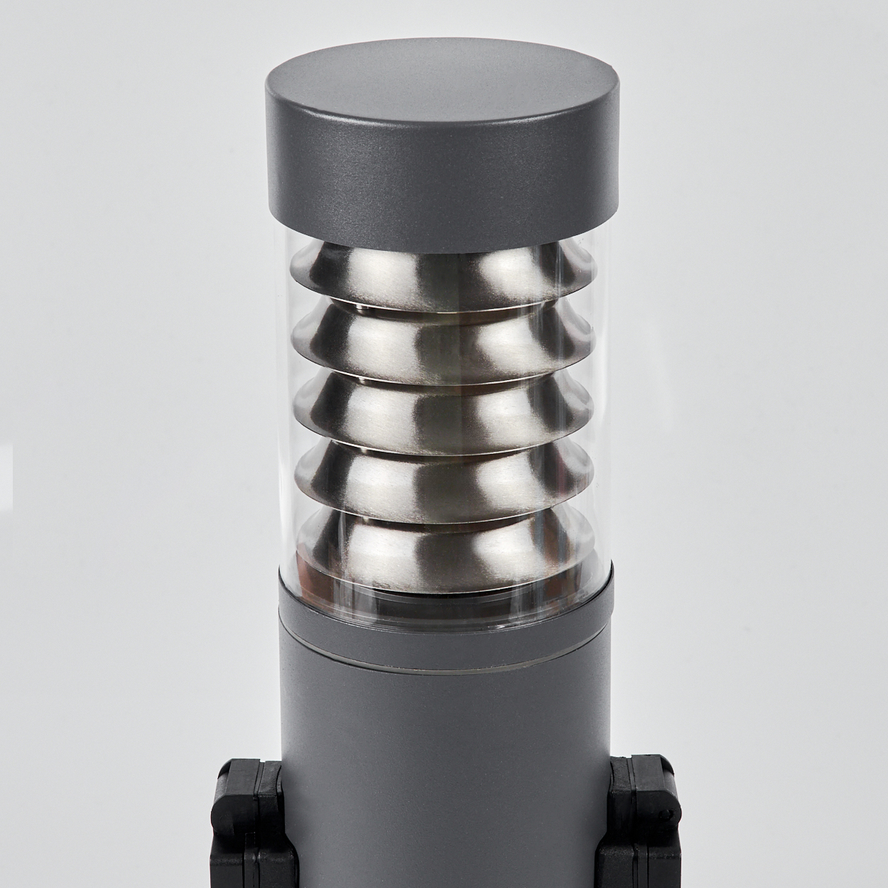 Dunkelgraue Außenlampe mit Metallplatten in einem Glasrohr, auf weißem Hintergrund.