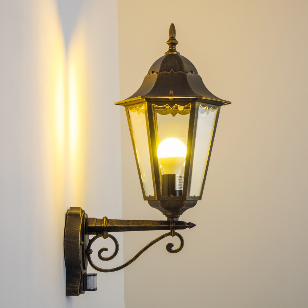Eine verzierte Außenwandleuchte mit Gold-Finish und klarem Glasschirm, beleuchtet mit einer LED-Glühbirne.