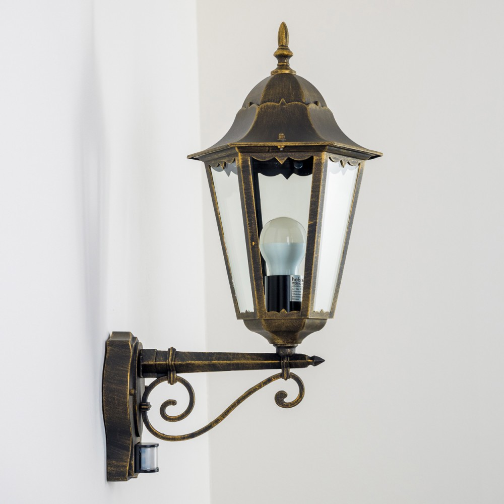 Eine verzierte Außenwandleuchte mit Gold-Finish und klarem Glasschirm, beleuchtet mit einer LED-Glühbirne.