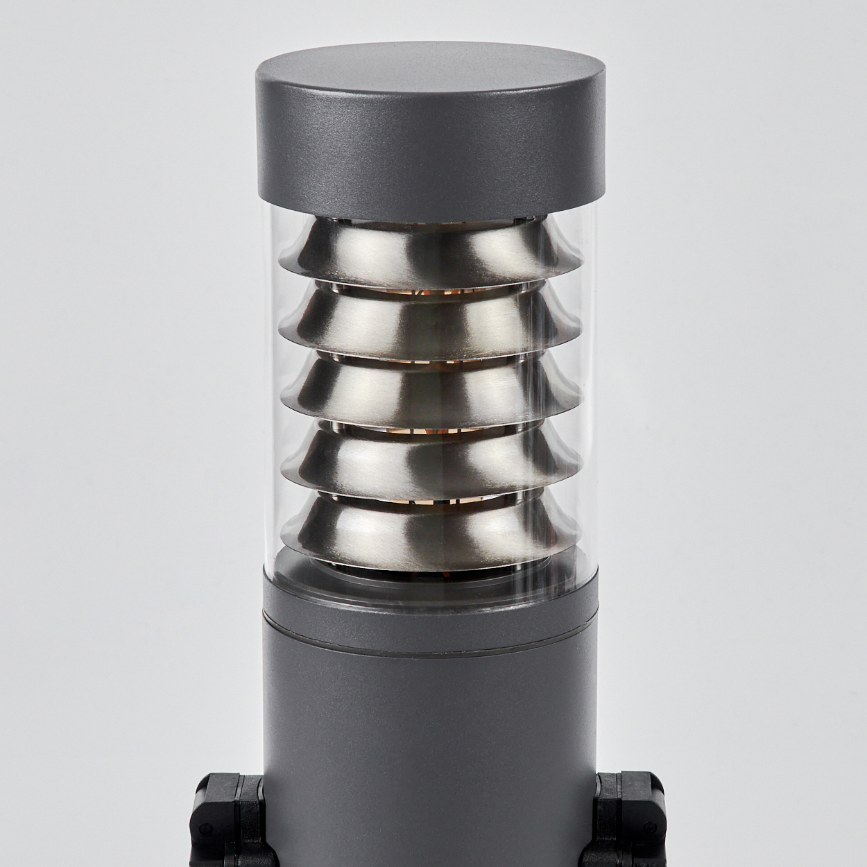 Nahaufnahme einer zylindrischen grauen Außenlampe mit silbernen horizontalen Lamellen in einer klaren Glasabdeckung.