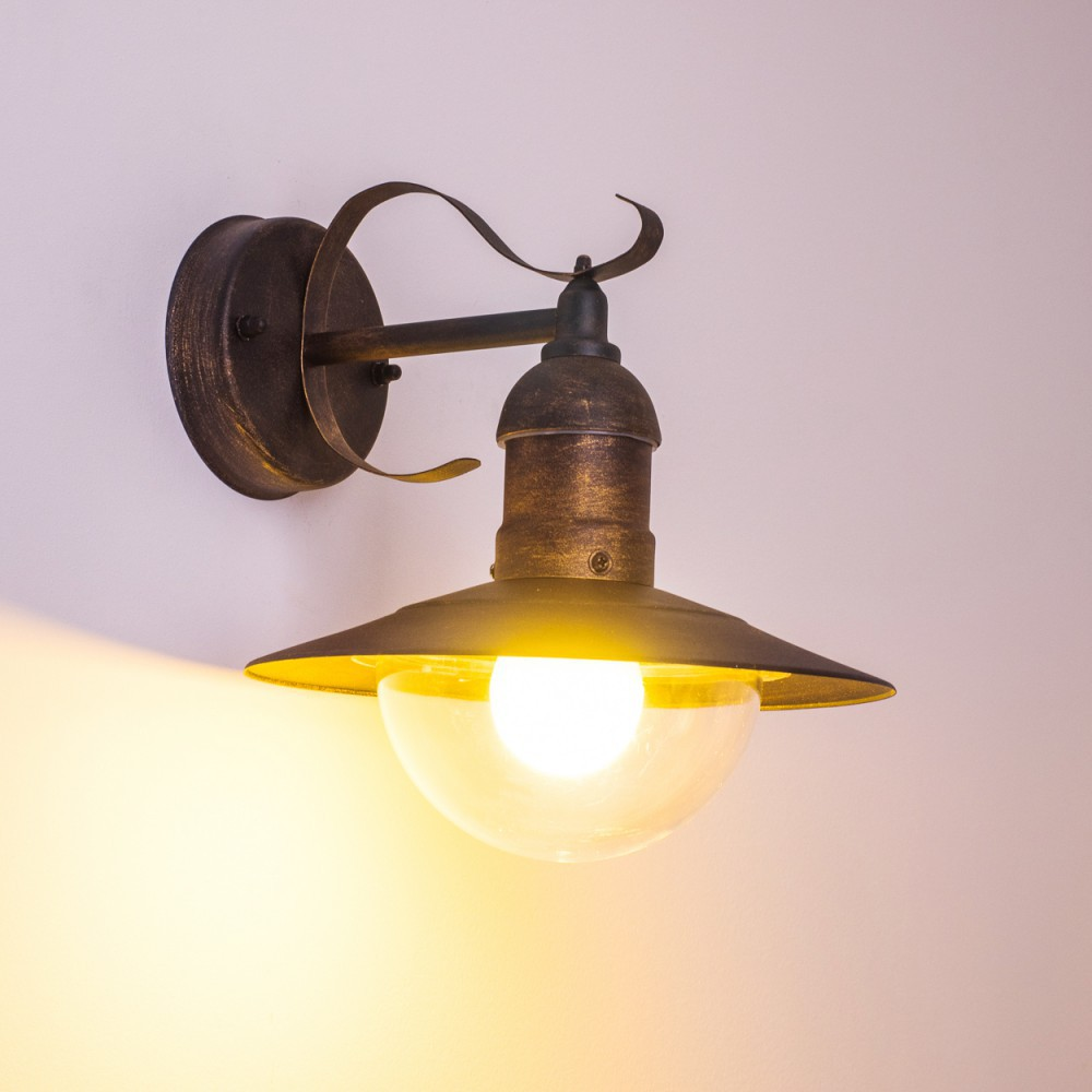 Bronzefarbene Außenwandleuchte mit klarem, halbkugelförmigem Schirm, beleuchtet mit warmem Licht. Weißer Hintergrund.