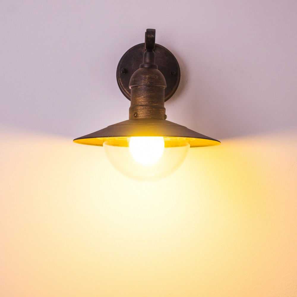 Bronzefarbene Außenwandleuchte mit klarem, halbkugelförmigem Schirm, beleuchtet mit warmem Licht. Weißer Hintergrund.