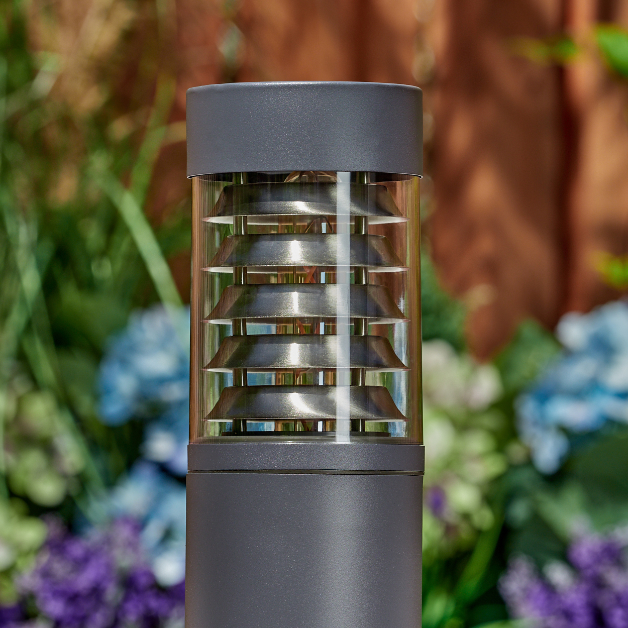 Nahaufnahme einer grauen Außenlampe. Sie hat einen klaren, geriffelten Glasaufsatz und steht im Garten.