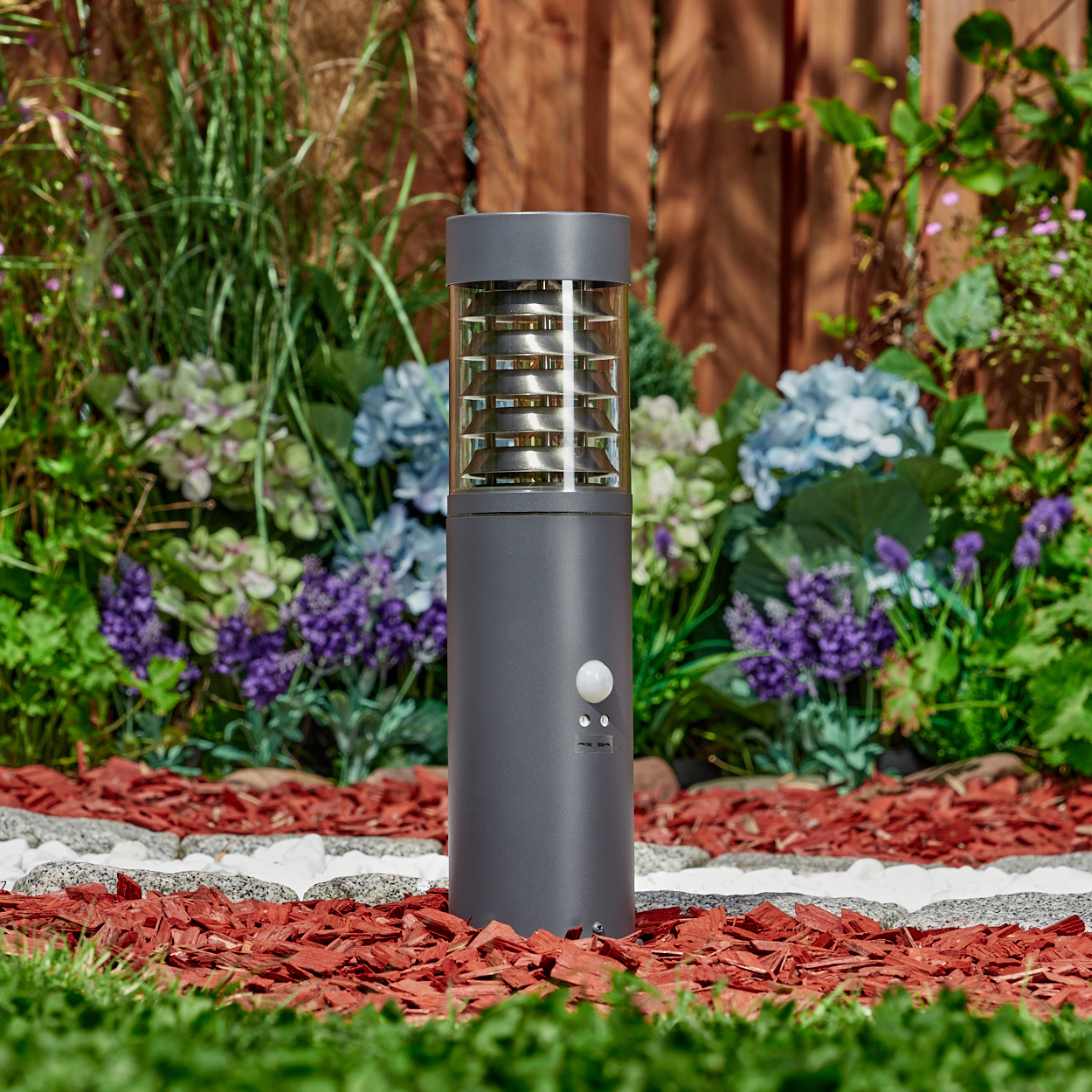 Eine graue Außenlampe mit Bewegungsmelder, umgeben von Grün und Blumen, in der Nähe eines Holzzauns.