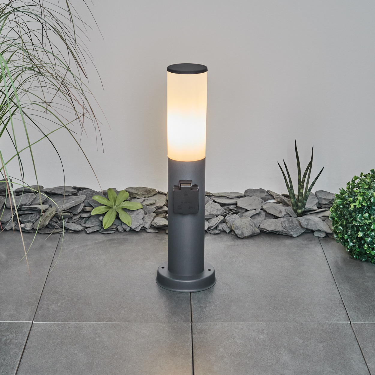 Graue Außenlampe mit beleuchtetem Oberteil. Busch- und Steinhintergrund, mit Licht an.