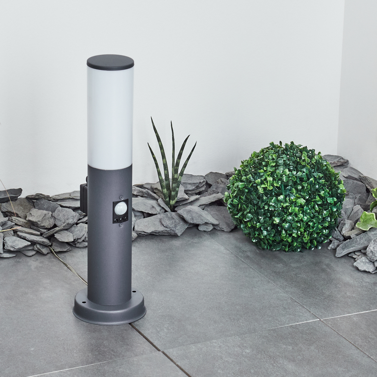 Graue Außenlampe mit grünem Busch- und Steinhintergrund neben einer kleinen Pflanze.