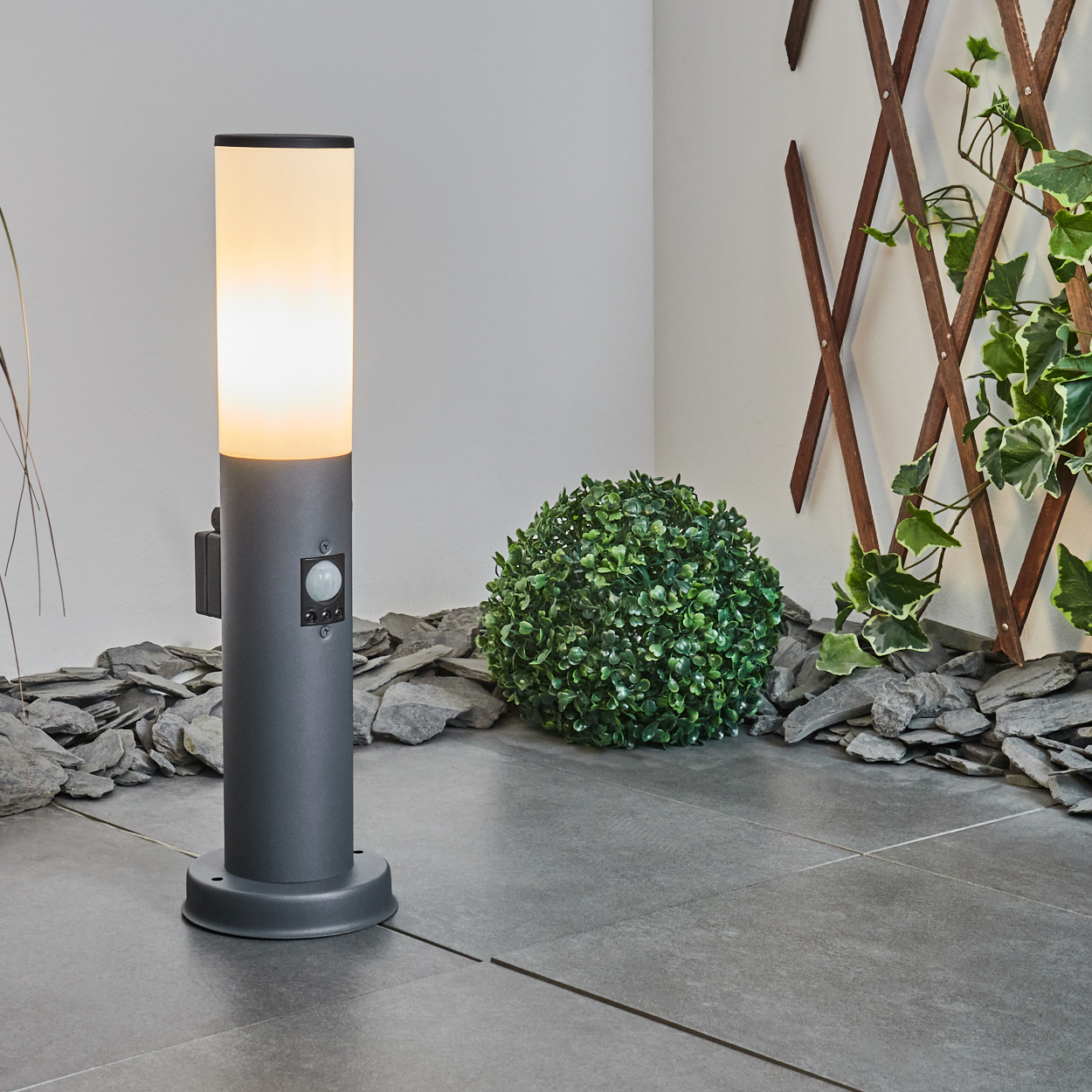 Graue, zylindrische Außenlampe mit weißem Oberteil, beleuchtet. Grüner Busch und Steine im Hintergrund.