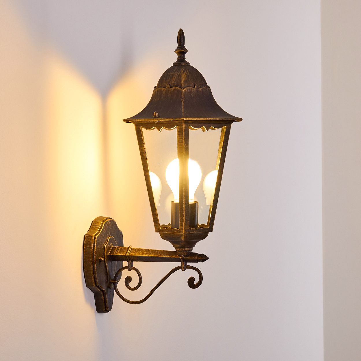 Verzierte Außenwandleuchte mit drei brennenden Glühbirnen, die ein warmes Licht auf eine helle Wand werfen.